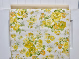 A 1960s vintage floral wallpaper with a retro yellow, orange, and green rose pattern on an off-white background, shown hanging on a white wooden wall.
